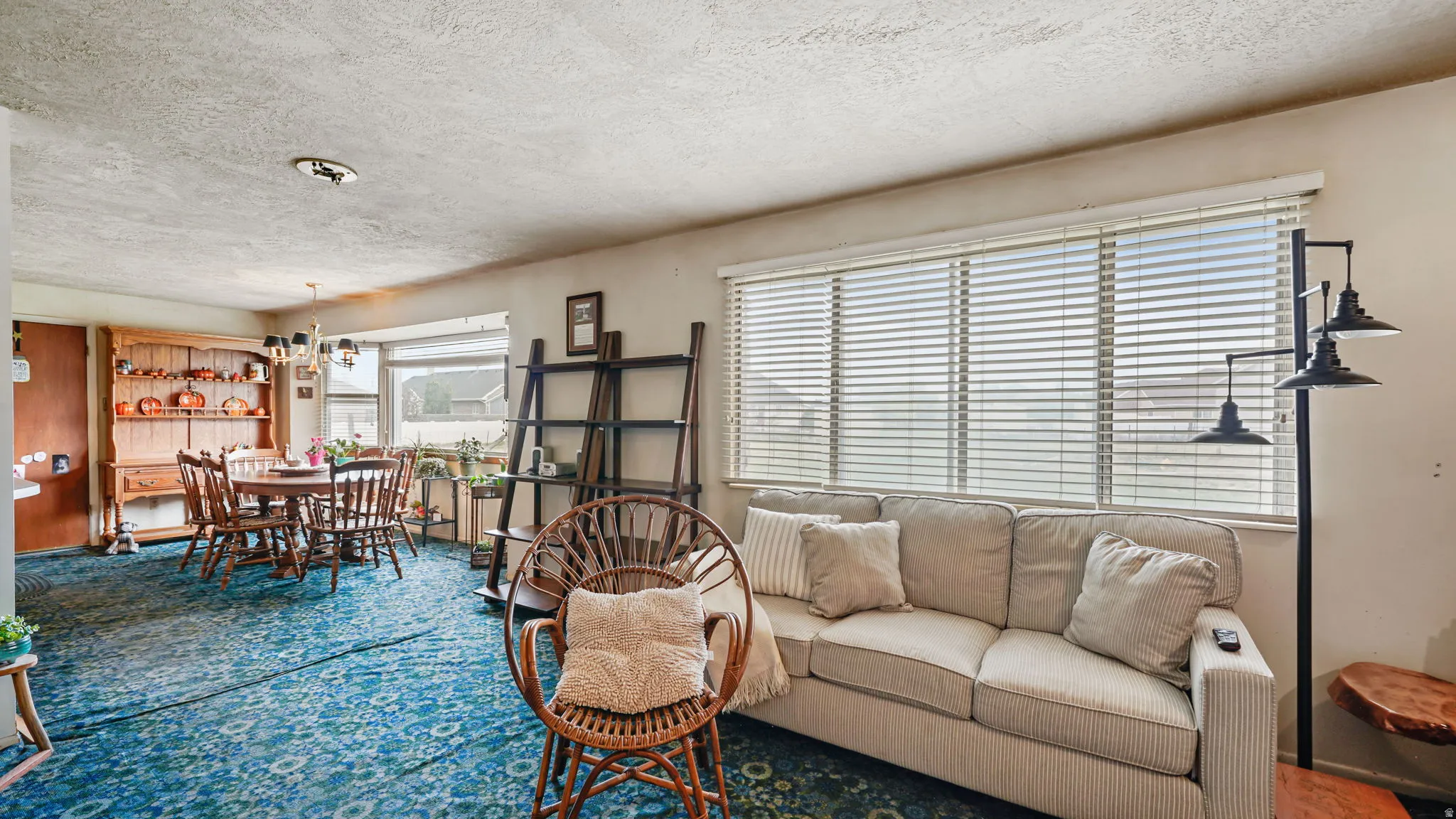 Spacious living area featuring patterned carpeting, large windows with horizontal blinds, and a dining area with a corner bay window