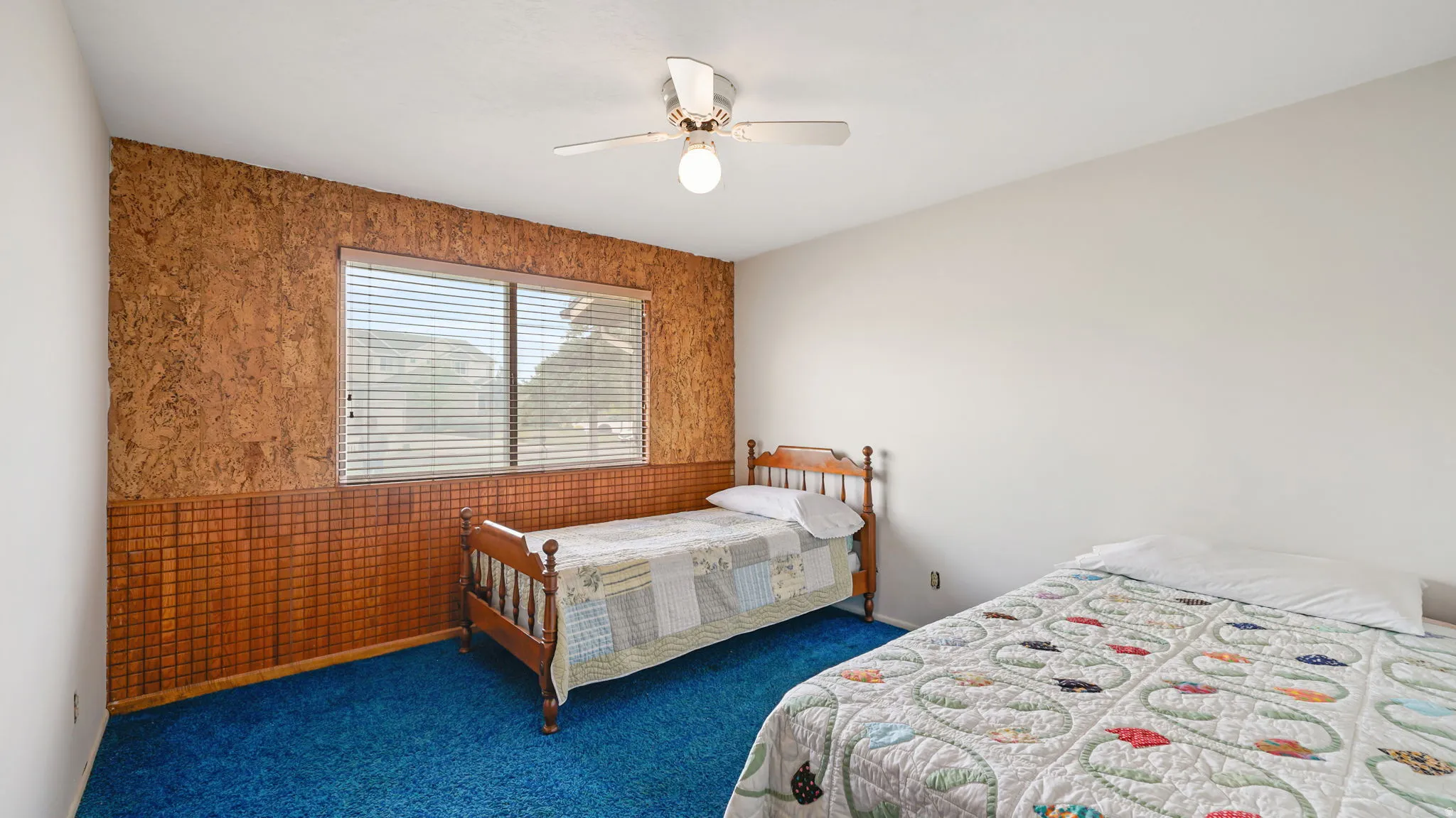 Interior room featuring a window with blinds, a ceiling fan with integrated lighting, textured wood-paneling on one wall, and blue carpeting