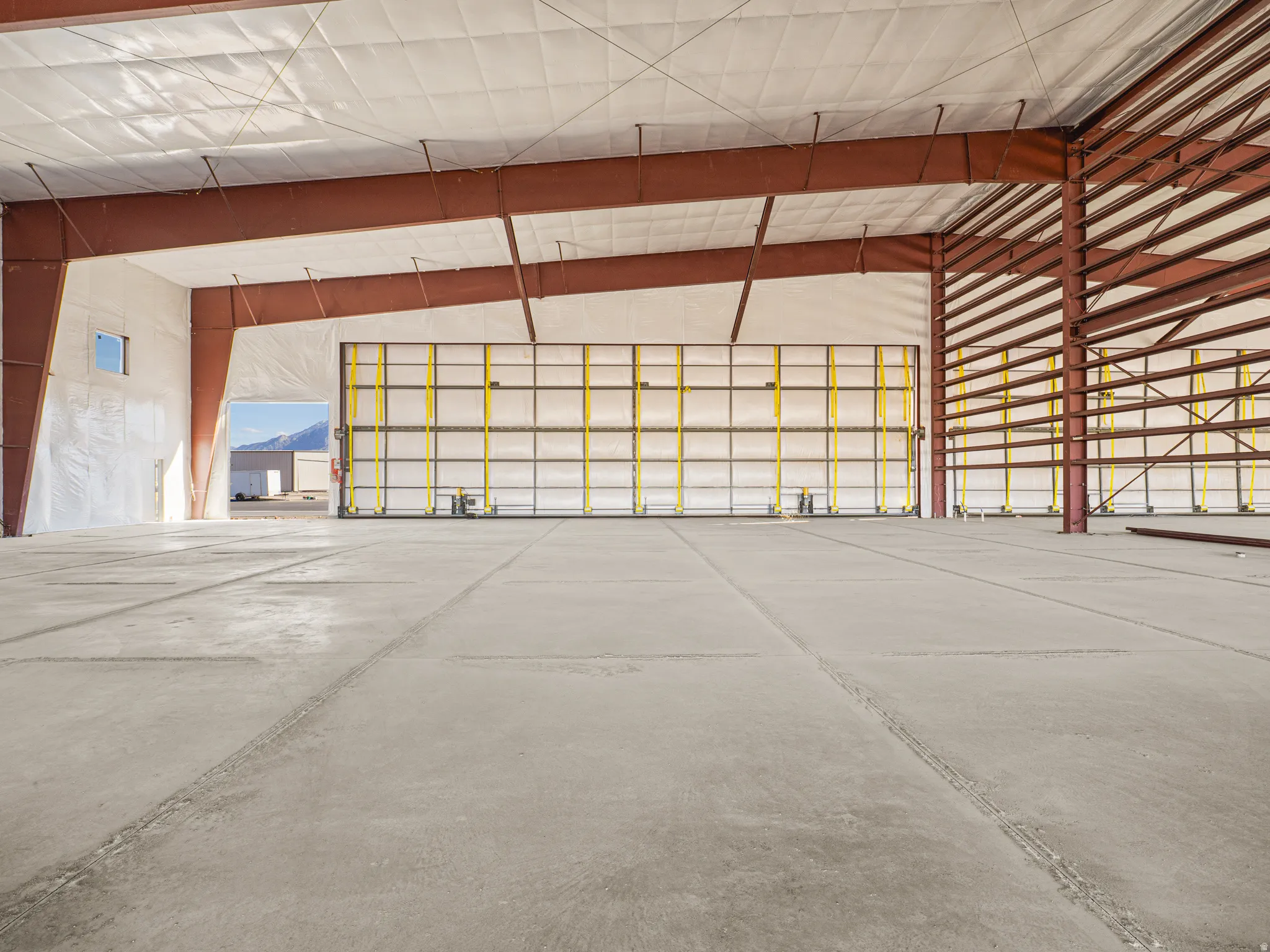 Expansive industrial interior featuring a large roll-up door, exposed red steel structural beams, insulated ceiling, and concrete flooring