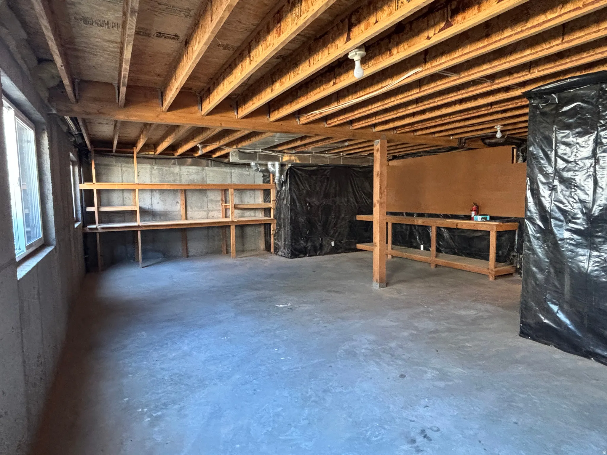 Unfinished basement featuring exposed wood beams, concrete foundation walls, two windows providing natural light, built-in shelving, and a large workbench