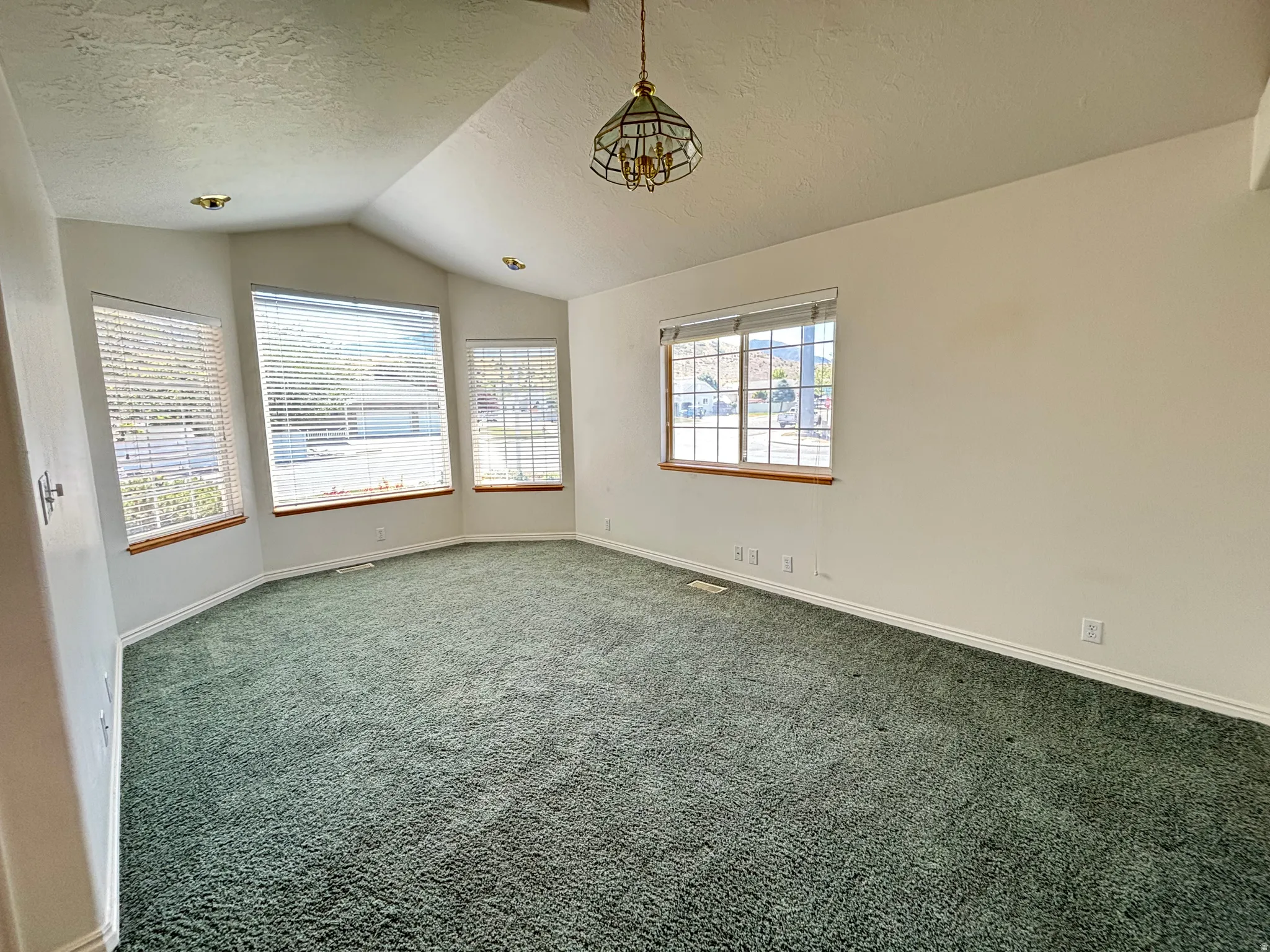 Spacious room featuring vaulted ceilings, a prominent bay window, green carpeting, white baseboards, and a central chandelier