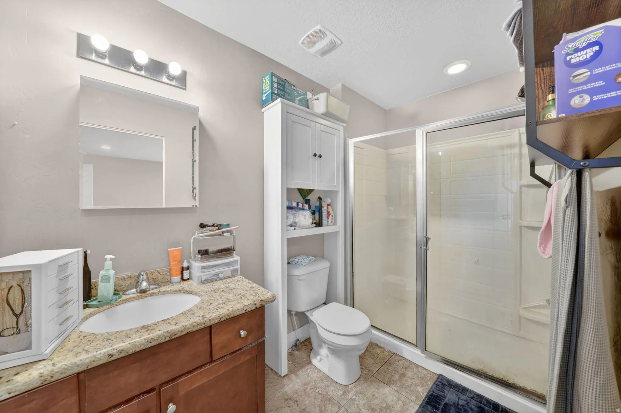 Full bathroom featuring a single vanity with a granite countertop and wood-finish cabinetry, a framed mirror, and a glass-enclosed shower with a built-in shelving unit