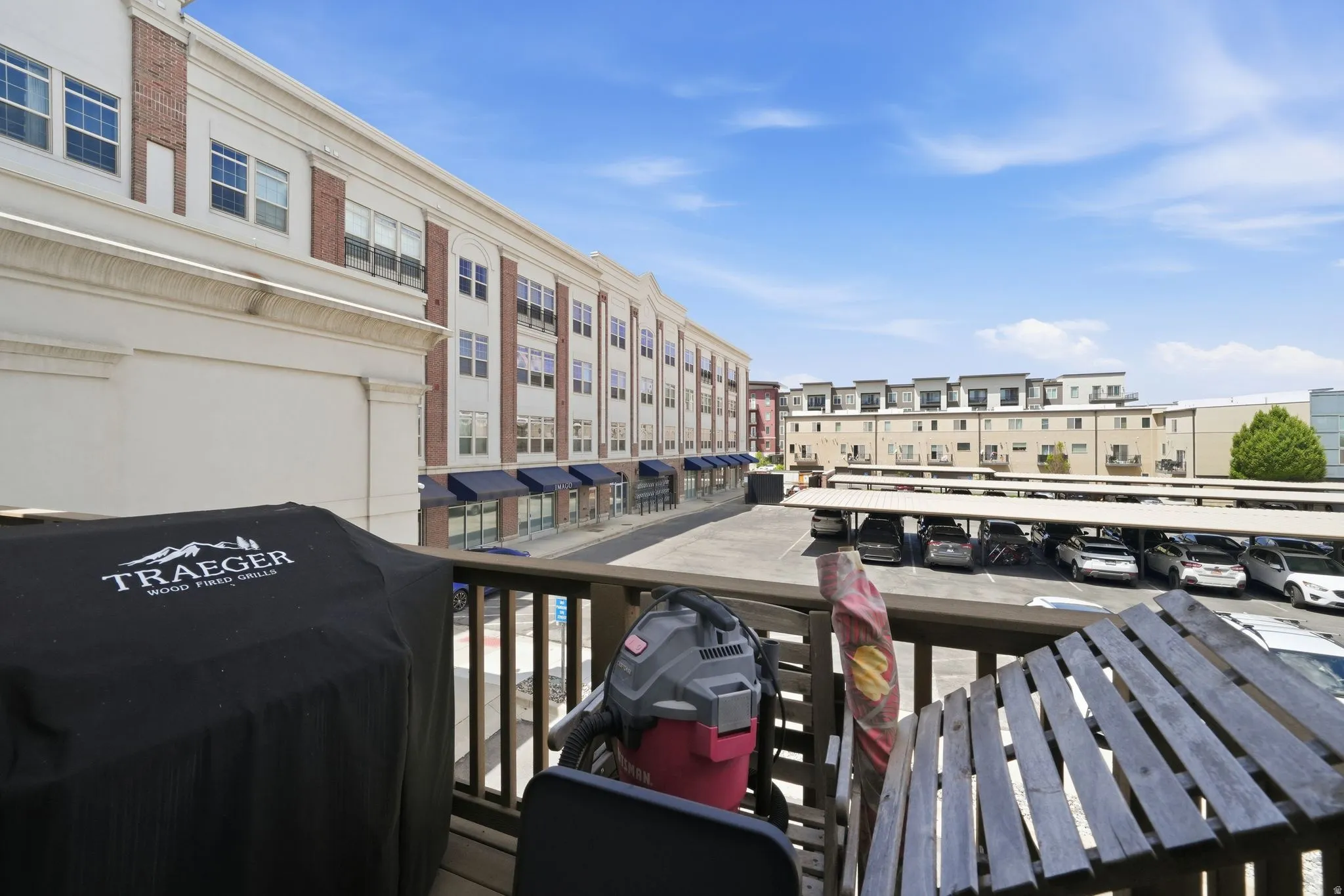 Balcony featuring wood railing and flooring, offering views of the surrounding complex with brick and white facade buildings and covered parking structures