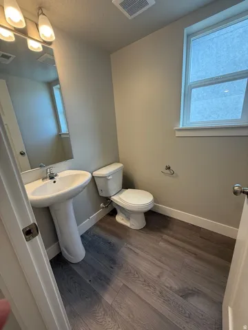 Bathroom featuring a pedestal sink, a toilet, wood-finish flooring, a rectangular window, and a wall-mounted vanity light fixture