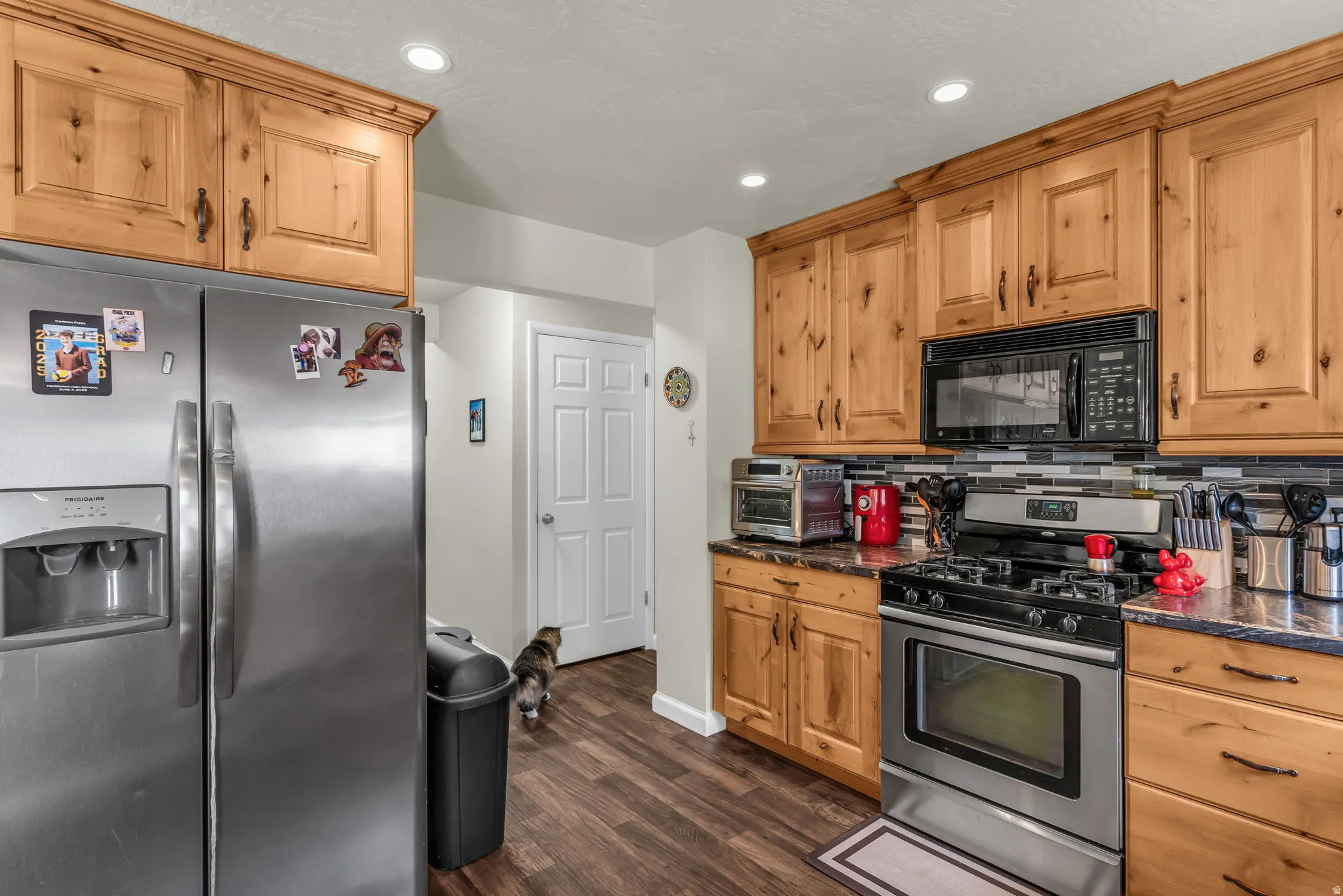 Kitchen featuring wood-finish cabinetry, stainless steel appliances, a black and gray mosaic tile backsplash, dark countertops, and wood-finish flooring