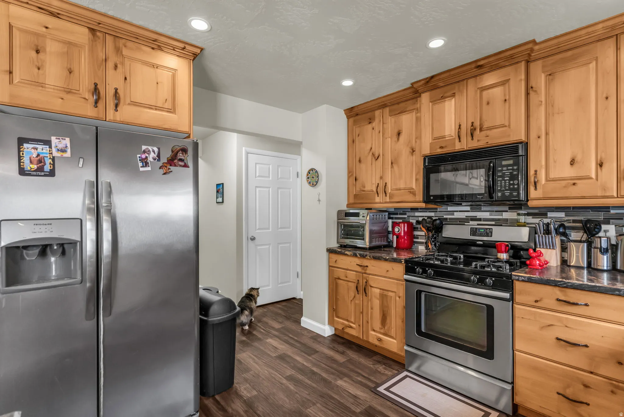 Kitchen featuring wood cabinetry, stainless steel appliances, dark countertops, a tile backsplash, and recessed lighting