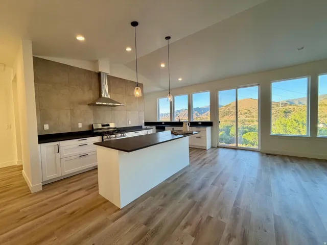 Open-concept kitchen featuring white cabinetry, dark countertops, a stainless steel range with a professional-grade hood, a central island, and wood-finish flooring, complemented by multiple large windows overlooking a mountainous landscape