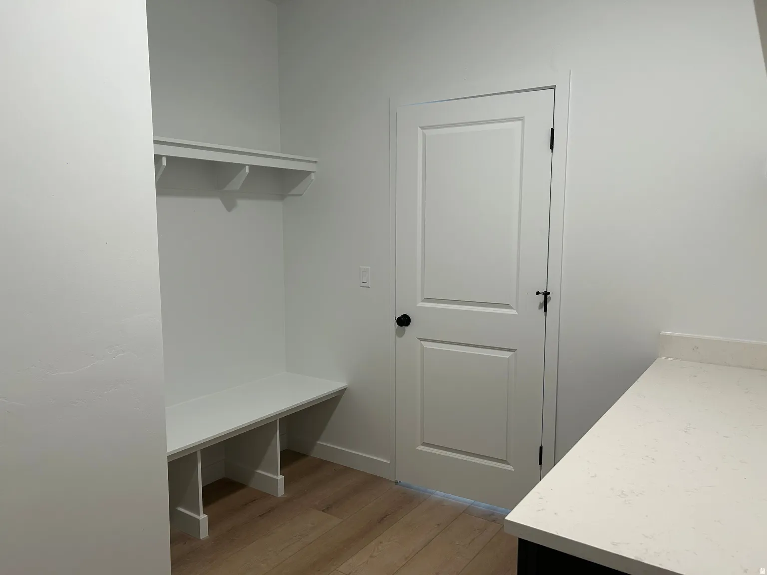Built-in organizational bench with shelving, wood-finish flooring, white paneled door with black hardware, and a light-toned countertop