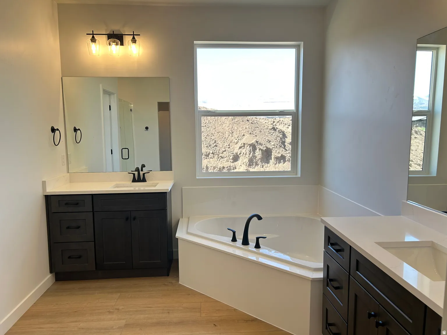 Spacious bathroom featuring an integrated corner soaking tub with matte black fixtures, dual dark wood vanities with white countertops, and wood-finish flooring