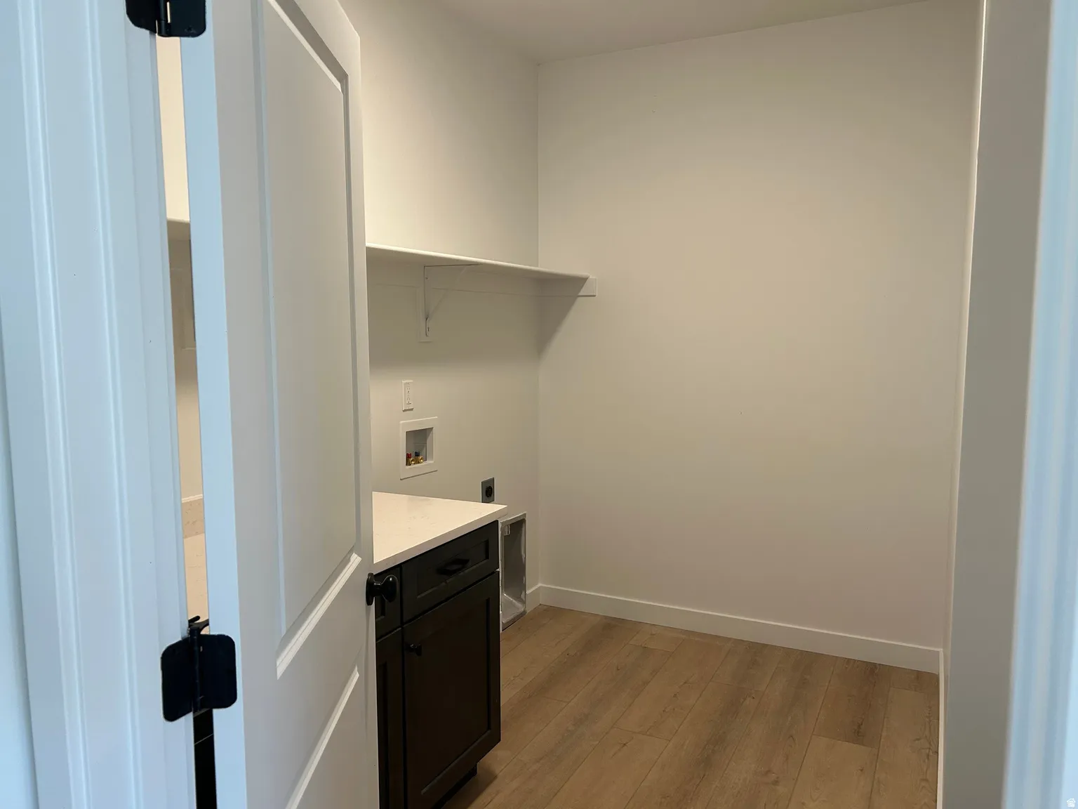 Laundry area featuring wood-finish flooring, a white wall shelf, a dark wood cabinet with a light-colored countertop, and visible utility hookups