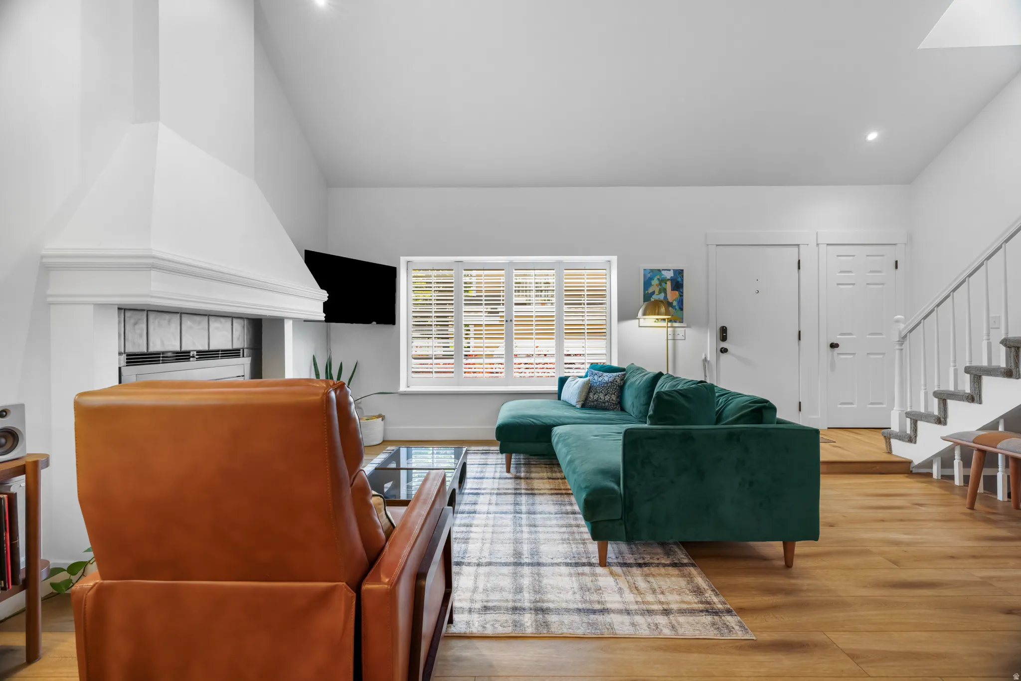Living area featuring vaulted ceilings, a built-in fireplace with a white mantel and tile surround, wood-finish flooring, recessed lighting, and a window with white louvered shutters