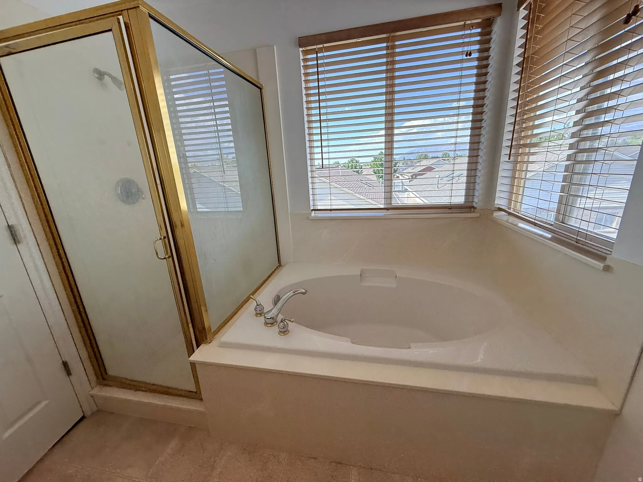 Bathroom featuring an oval built-in soaking tub with deck-mounted faucet, a separate glass-enclosed shower with gold-tone framing, and two windows with wood-finish blinds