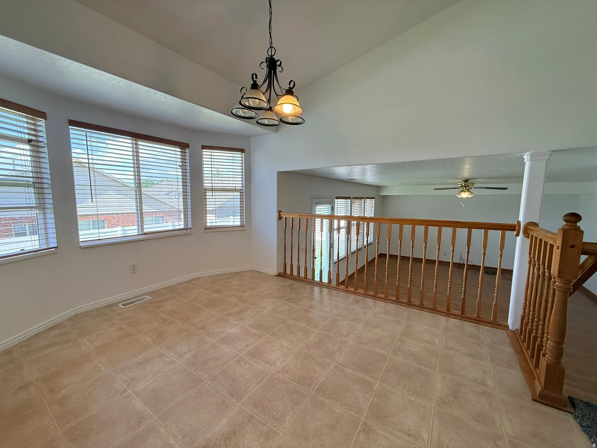 Vaulted ceiling with a chandelier light fixture, multiple windows with blinds, tiled flooring, wood railing, and a view to the lower level with wood-finish flooring