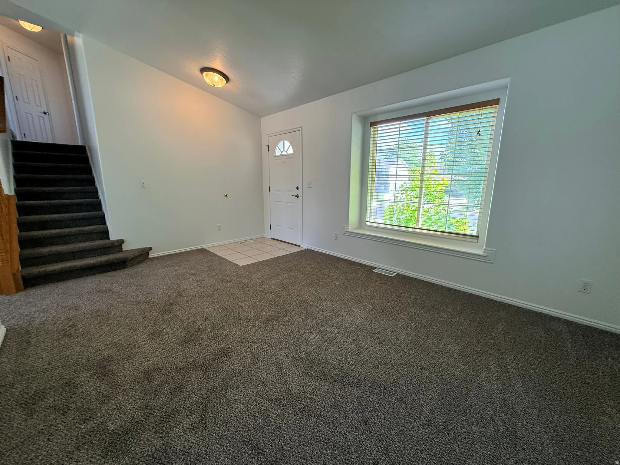 Spacious entryway featuring a large window, neutral wall tones, plush carpeting, a tiled entry area, and a staircase with wood railing