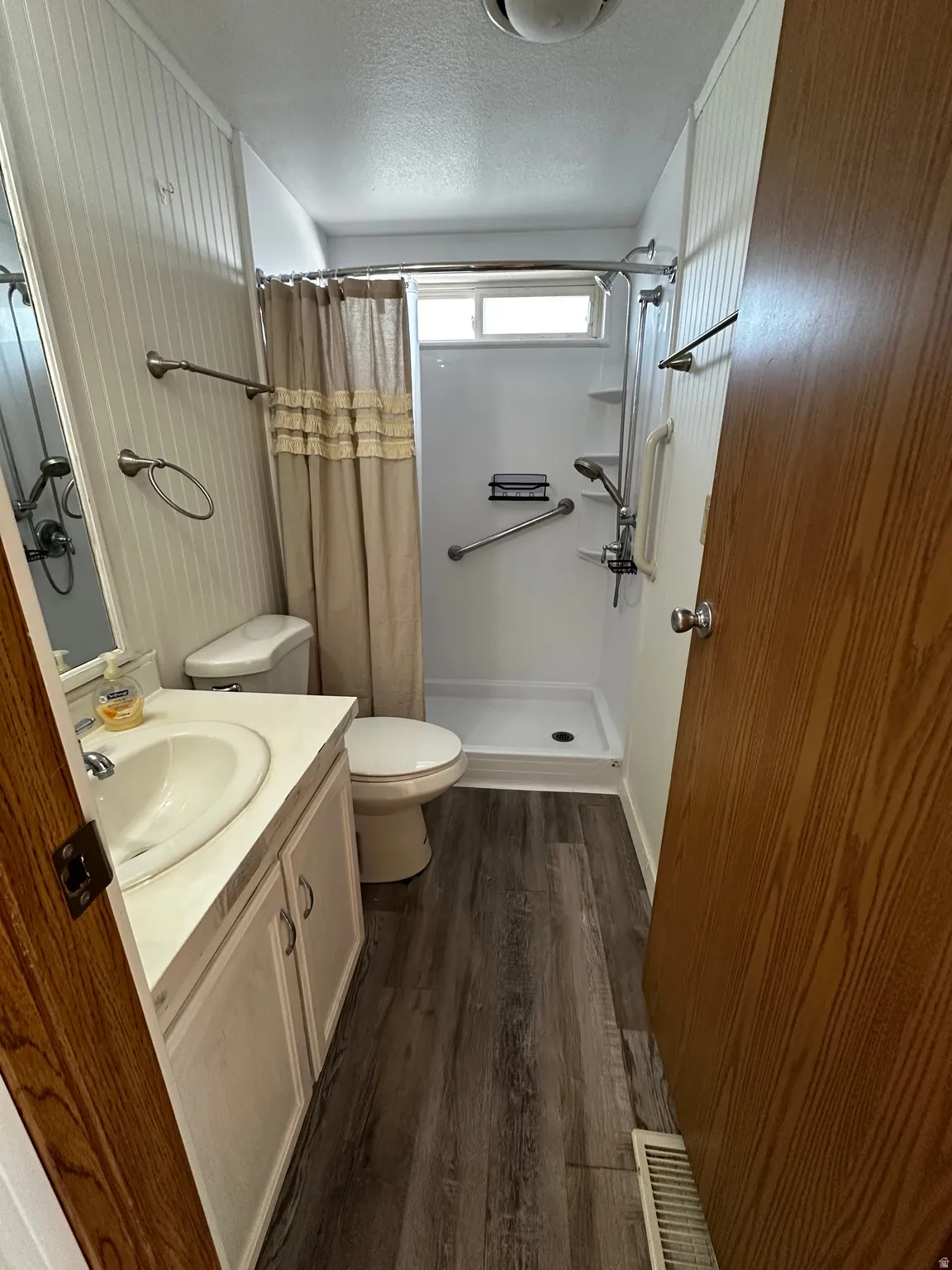 Bathroom featuring a walk-in shower with grab bars, a single vanity with storage, wood-finish flooring, and white paneled walls