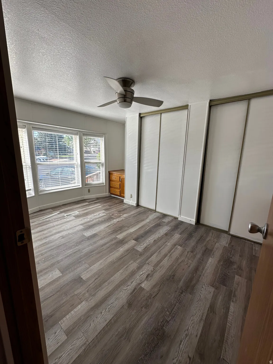 Spacious room featuring wood-finish flooring, a contemporary ceiling fan, and large windows with horizontal blinds