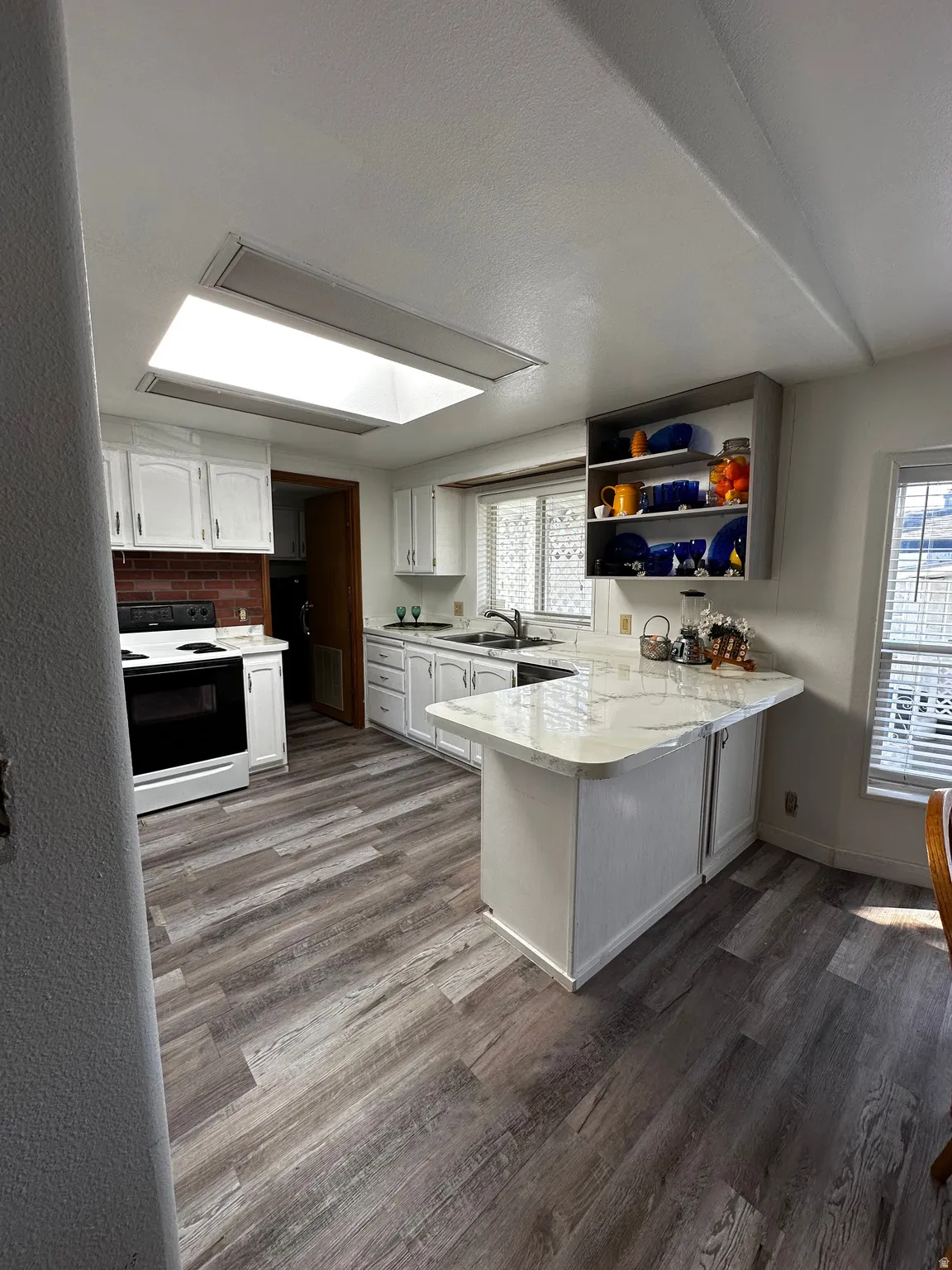 Spacious kitchen featuring wood-finish flooring, white cabinetry, a stone-finish countertop with an eat-at bar, a skylight, and a brick-finish backsplash