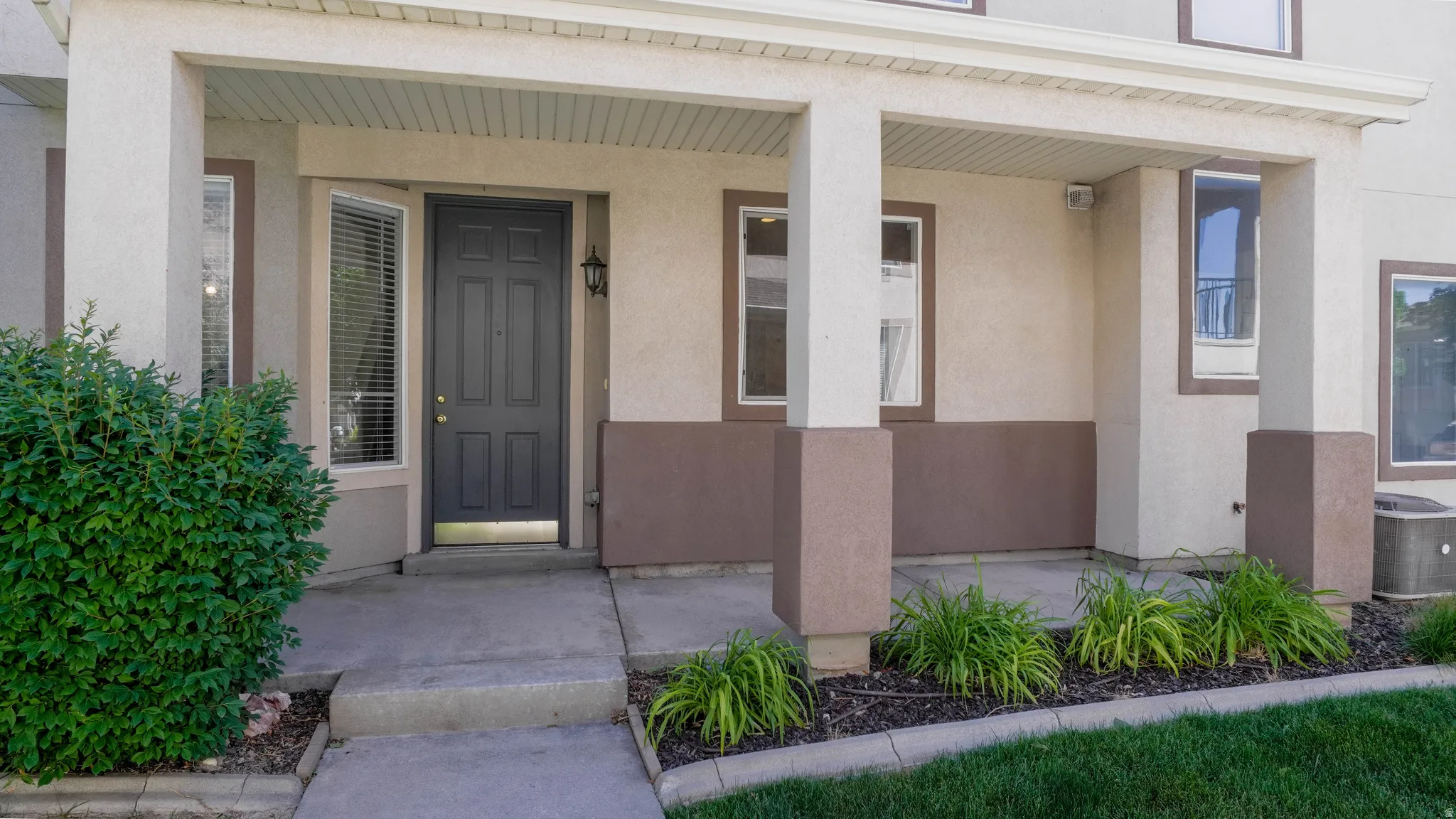 Covered front entry featuring a paneled door with sidelight, concrete porch, stucco exterior, and accent shrubbery