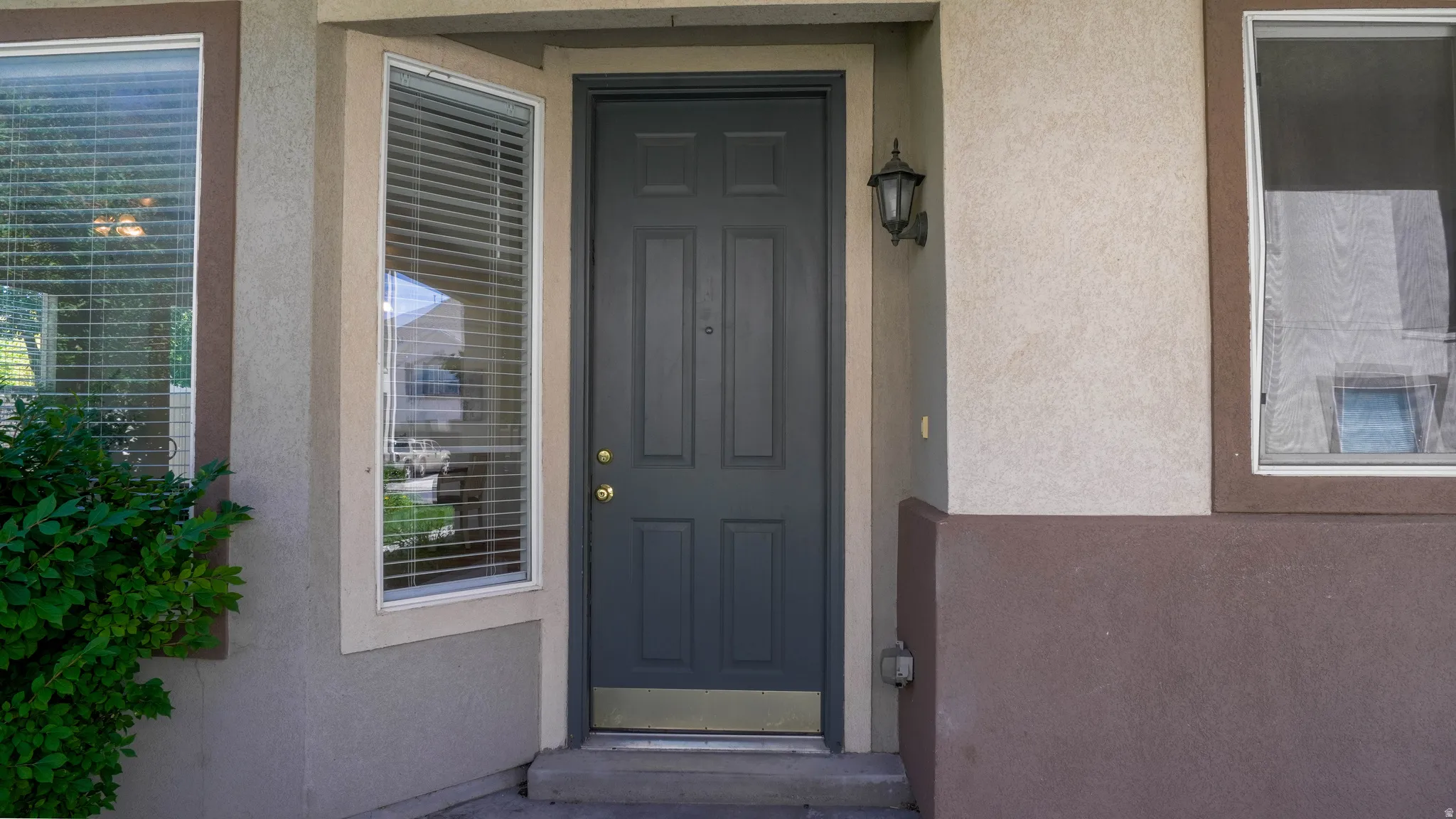 Exterior entryway featuring a paneled door, decorative wall sconce, bay window with blinds, and stucco siding