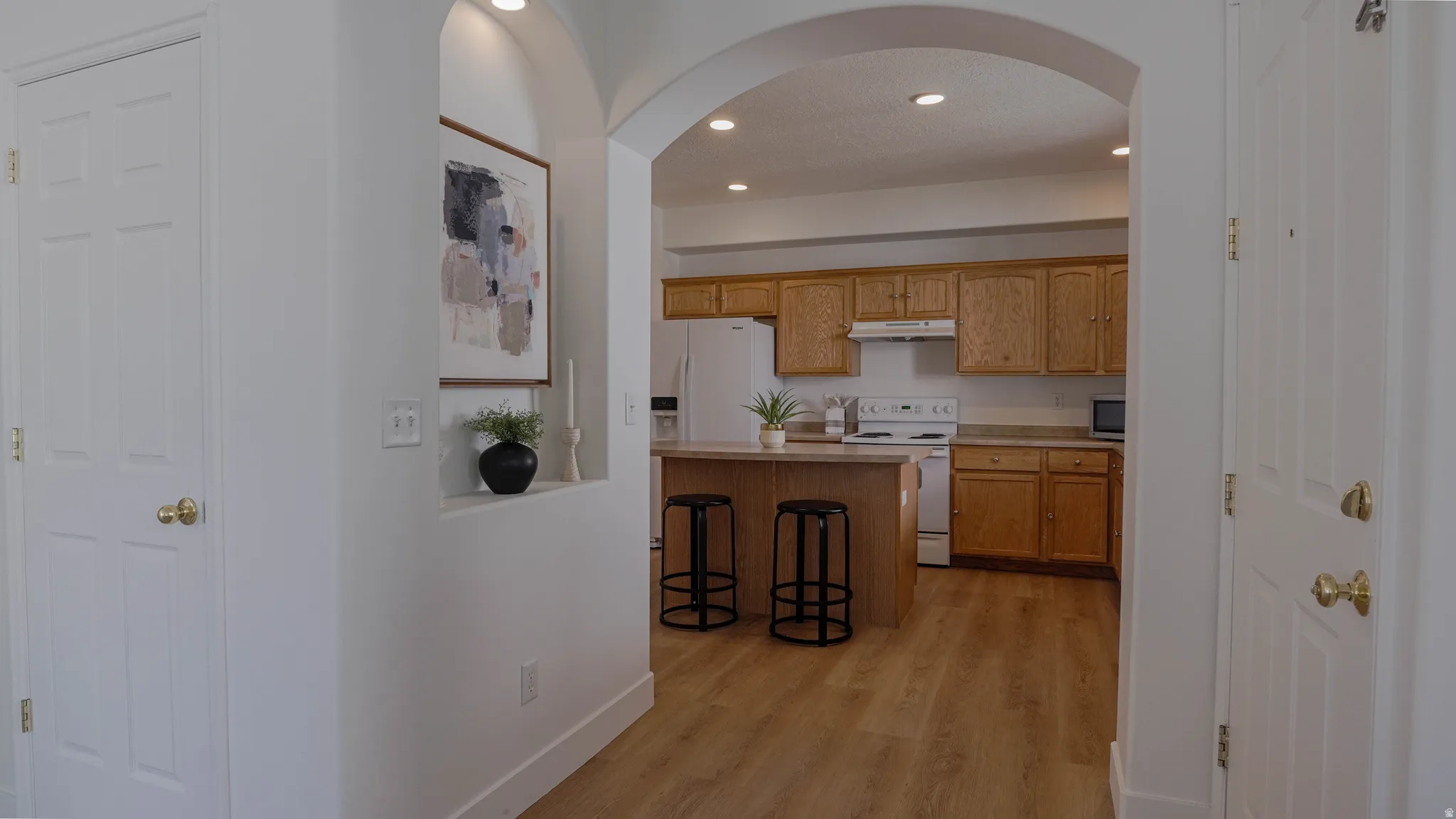 Arched entryway leading to a kitchen featuring wood-finish flooring, light wood cabinetry, white appliances, and recessed lighting