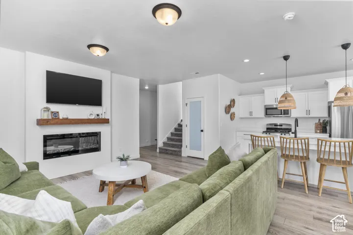 Open-concept living space featuring wood-finish flooring, a contemporary fireplace with a floating wood mantel, and an adjacent kitchen with white cabinetry and stainless steel appliances