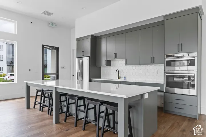 Contemporary kitchen featuring a large central island with seating, grey flat-panel cabinetry, stainless steel appliances, a hexagonal tile backsplash, and wood-finish flooring
