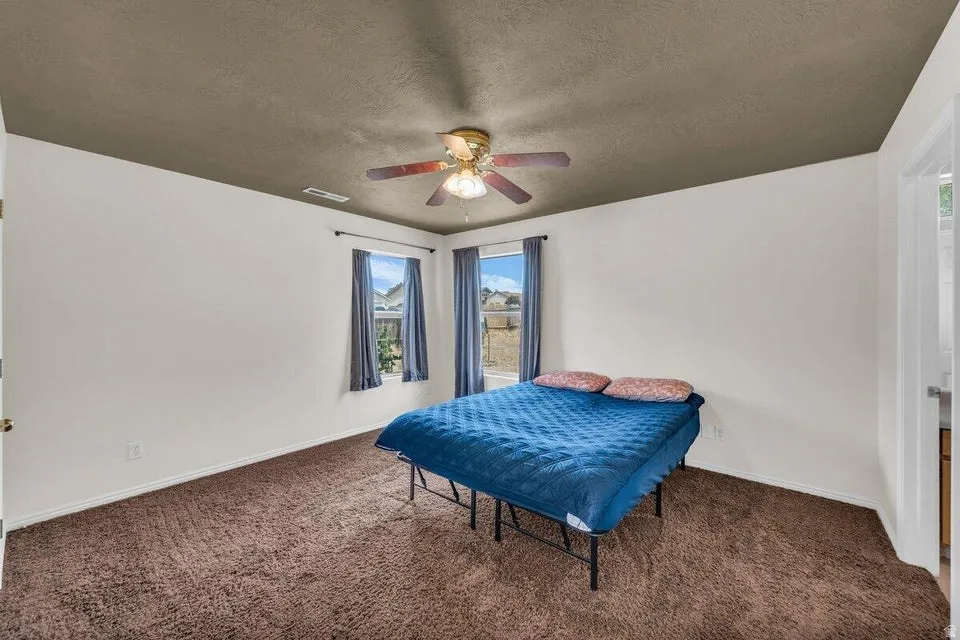 Bedroom featuring brown plush carpet, light-colored walls, a brown ceiling, a ceiling fan with light fixture, and two windows with dark curtains