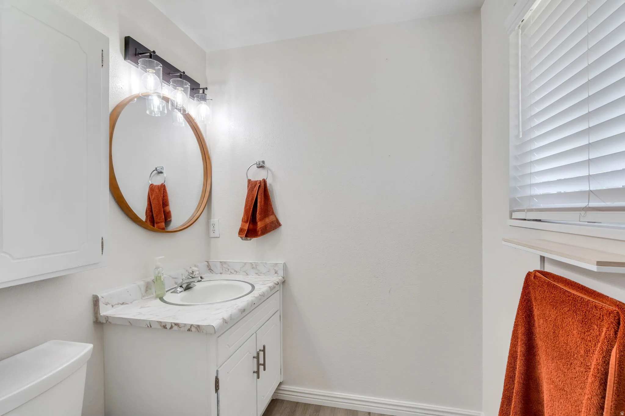 Primary bathroom vanity featuring white cabinets, light colored countertop, large mirror, window with blinds and vanity light fixture