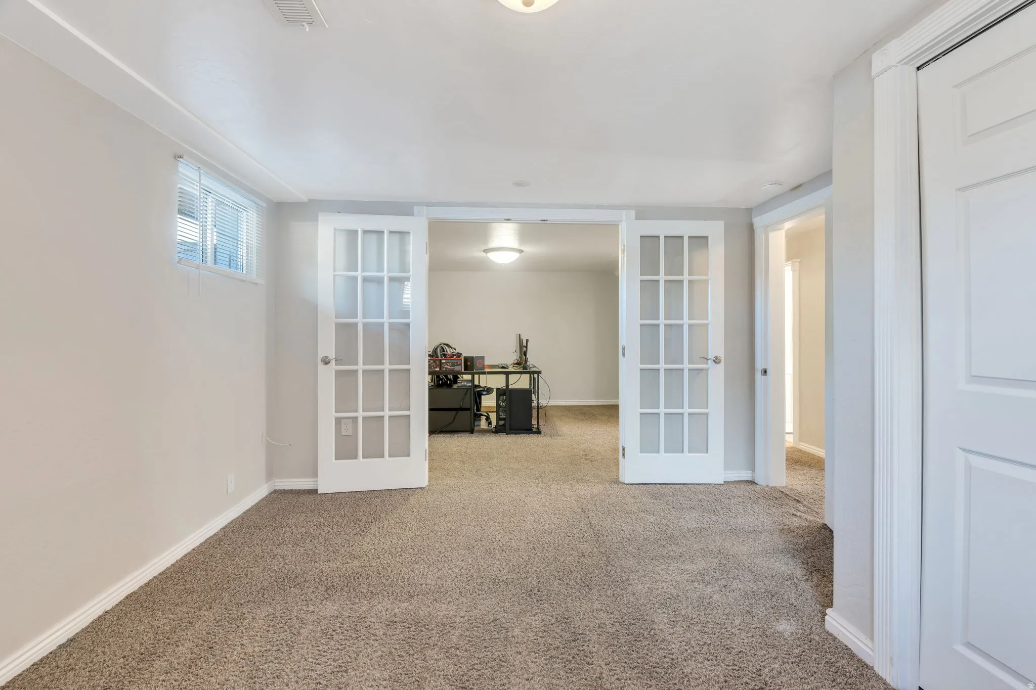 Apartment bedroom #2 with neutral-toned carpeting, two windows and a ceiling-mounted light fixture with twin French doors and white trim throughout