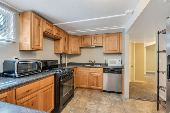 Apartment kitchen featuring natural wood-finish cabinetry, dark countertops, a stainless steel dishwasher, freestanding range, microwave and a window with blinds