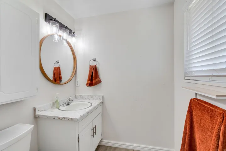 Primary bathroom vanity featuring white cabinets, light colored countertop, large mirror, window with blinds and vanity light fixture