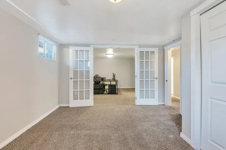 Apartment bedroom #2 with neutral-toned carpeting, two windows and a ceiling-mounted light fixture with twin French doors and white trim throughout