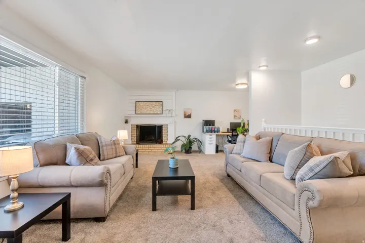 Carpeted living room featuring a brick fireplace with a white mantel, large window with blinds