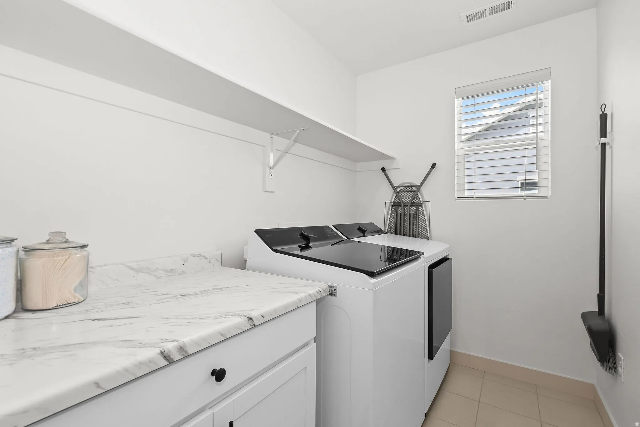 Dedicated laundry space featuring white cabinetry with black hardware, a light-toned countertop with veining, an upper storage shelf, and light tile flooring