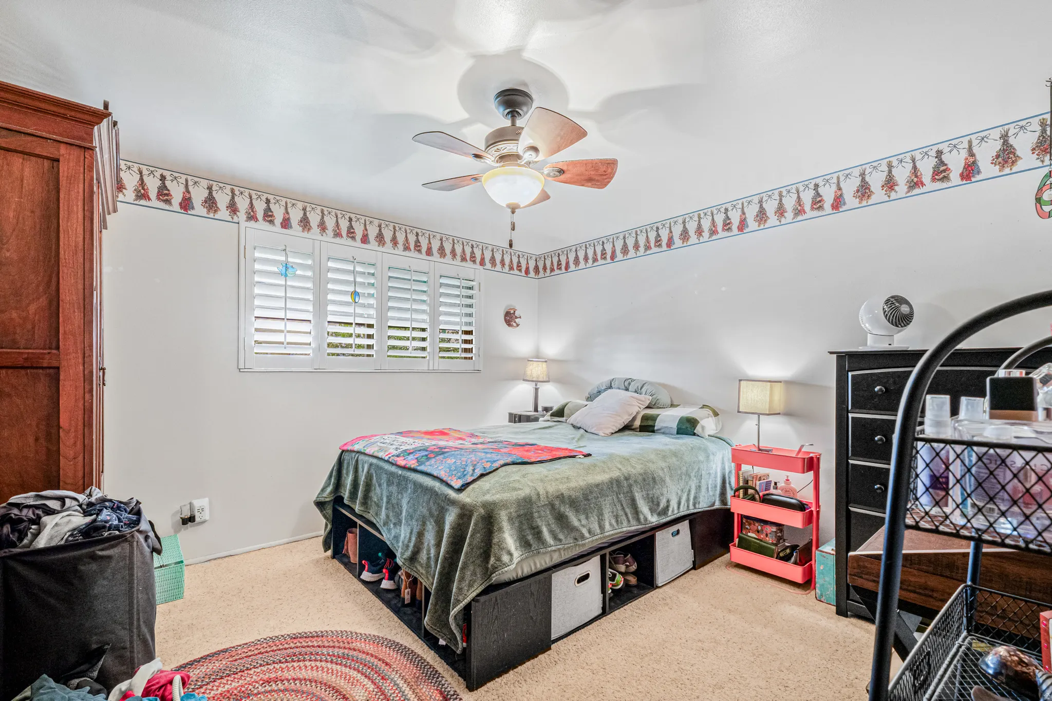 Bedroom featuring a ceiling fan with light fixture, carpeted flooring, and a window with plantation shutters