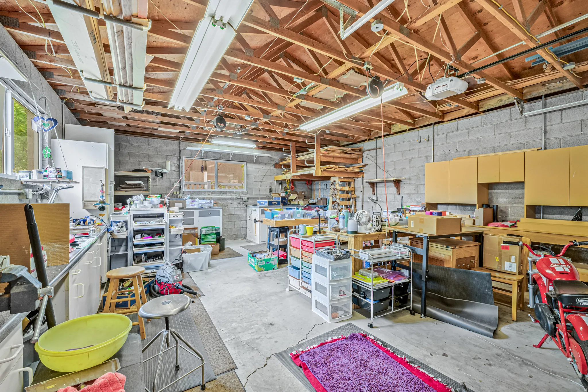 Spacious workshop area featuring exposed ceiling joists, concrete block walls, multiple windows for natural light, fluorescent lighting fixtures, and a concrete floor