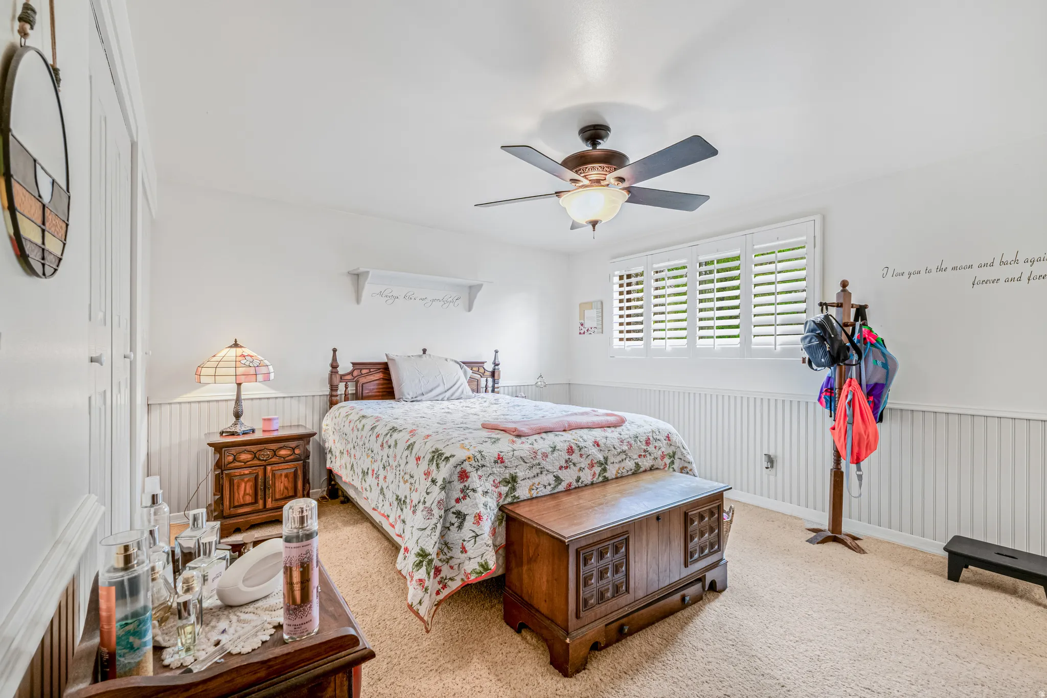 Carpeted room featuring a ceiling fan with integrated lighting, a window with white plantation shutters, and white vertical wainscoting