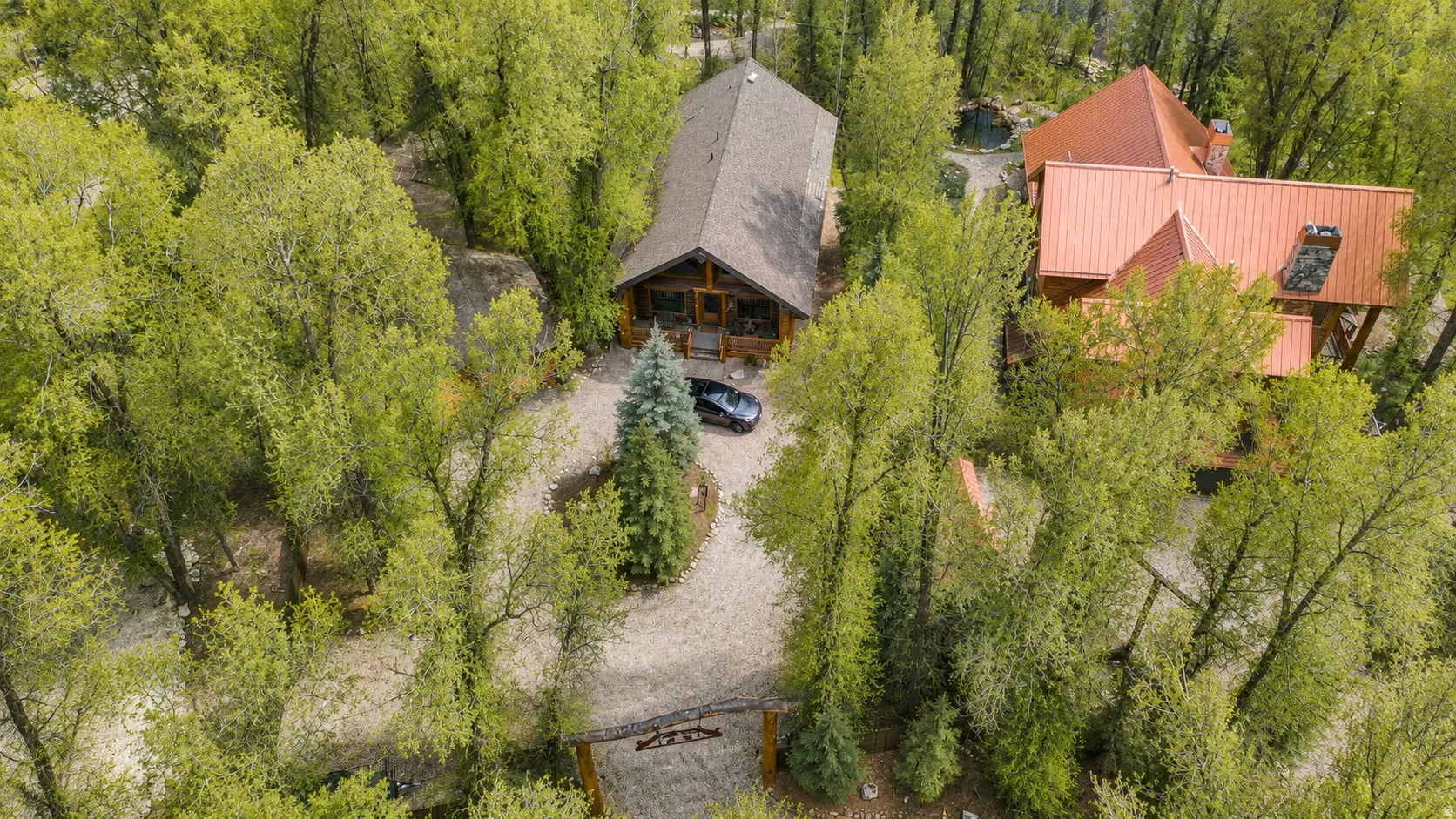 Secluded property featuring a primary residence with a dark roof, a secondary structure with a red metal roof, and a gravel driveway