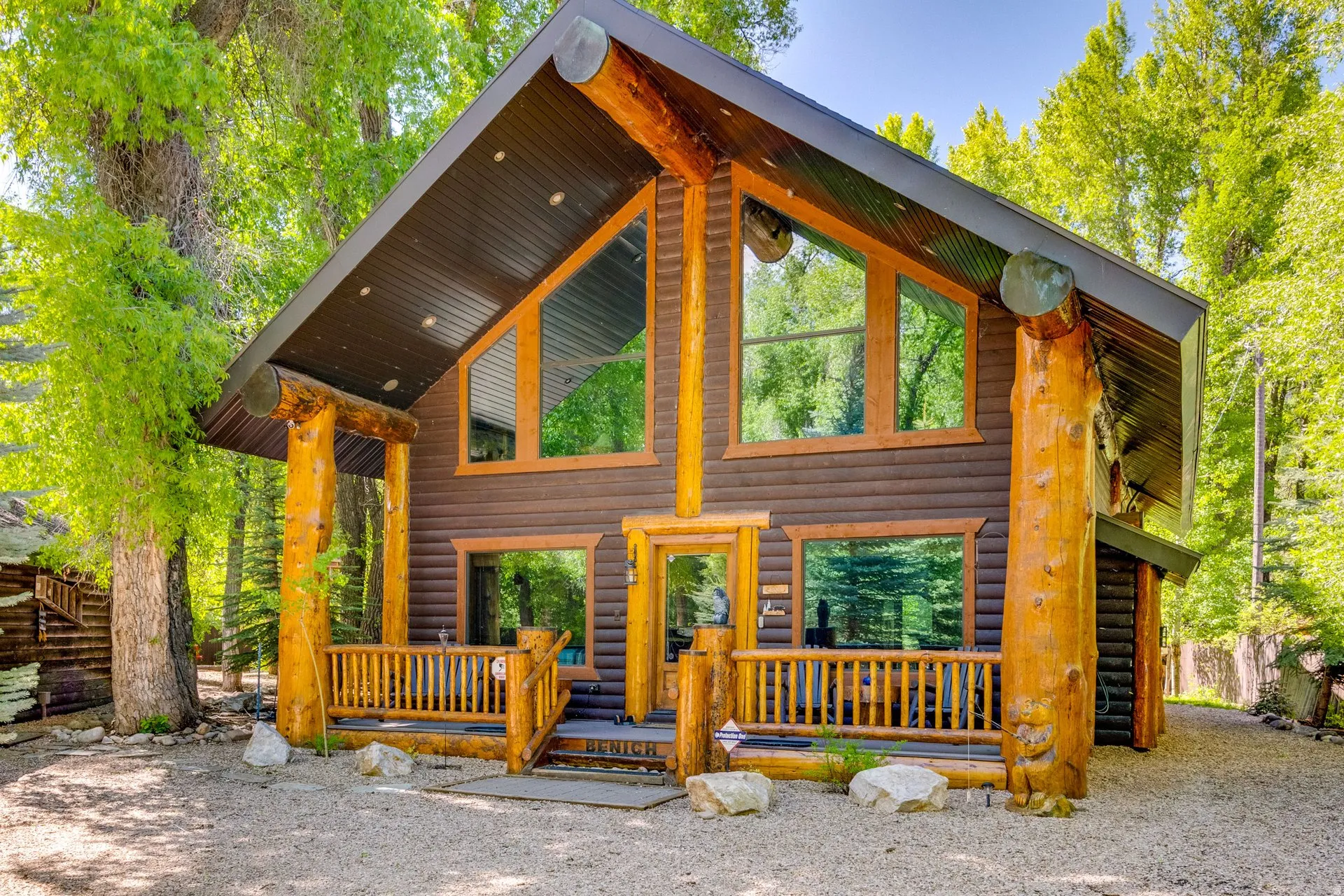 Log cabin architecture featuring prominent timber posts, expansive windows, a covered front porch with a wood railing, and a dark-toned corrugated metal roof