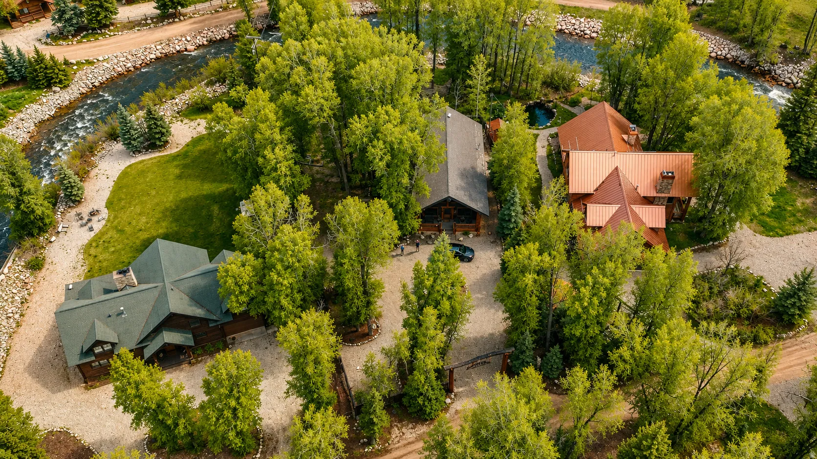 Lush property featuring three distinct structures, mature trees, a meandering river, expansive green lawns, and gravel pathways