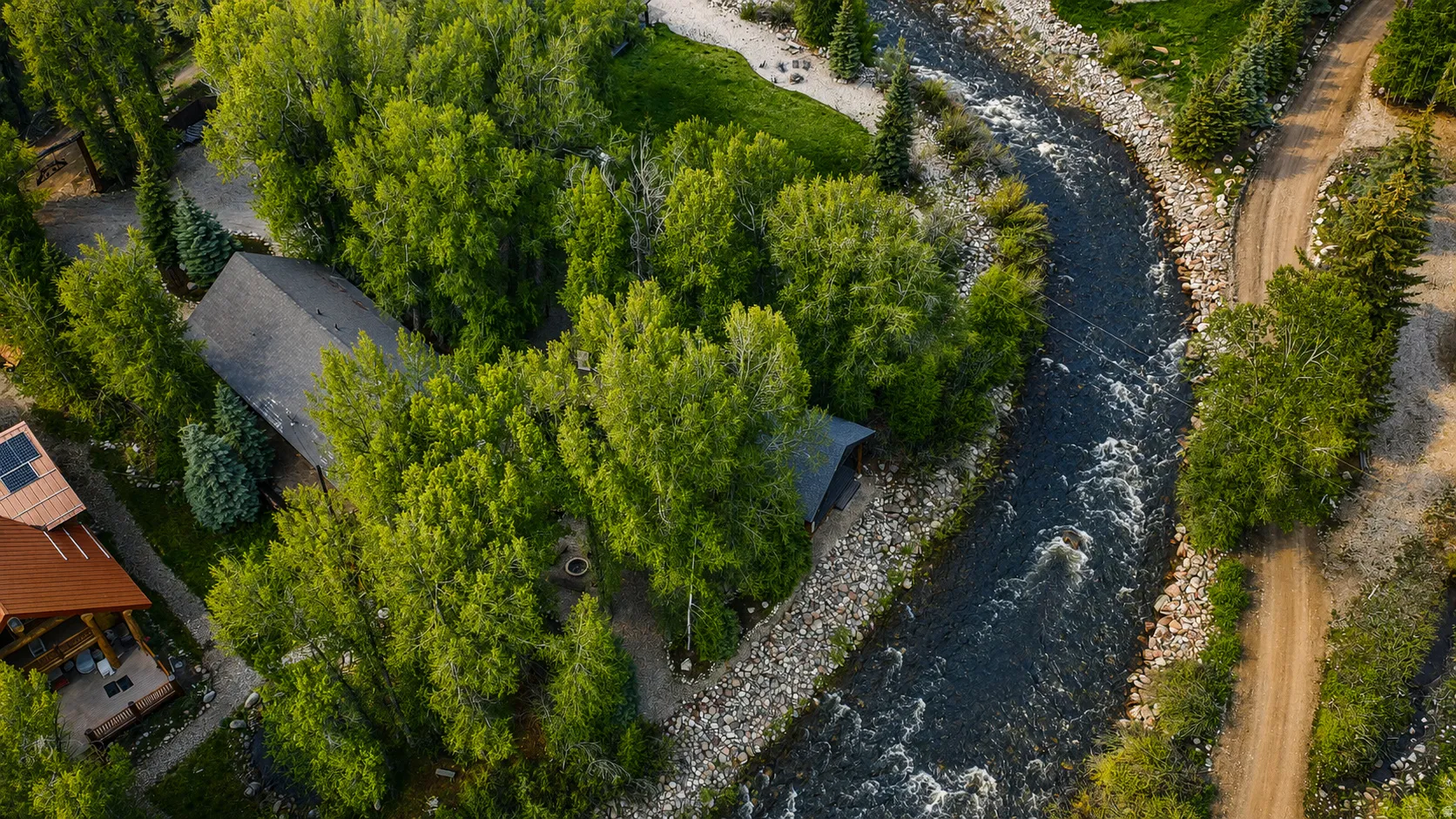 Riverside property featuring a pitched roof structure surrounded by mature trees, a natural stone riverbank, and a dirt path