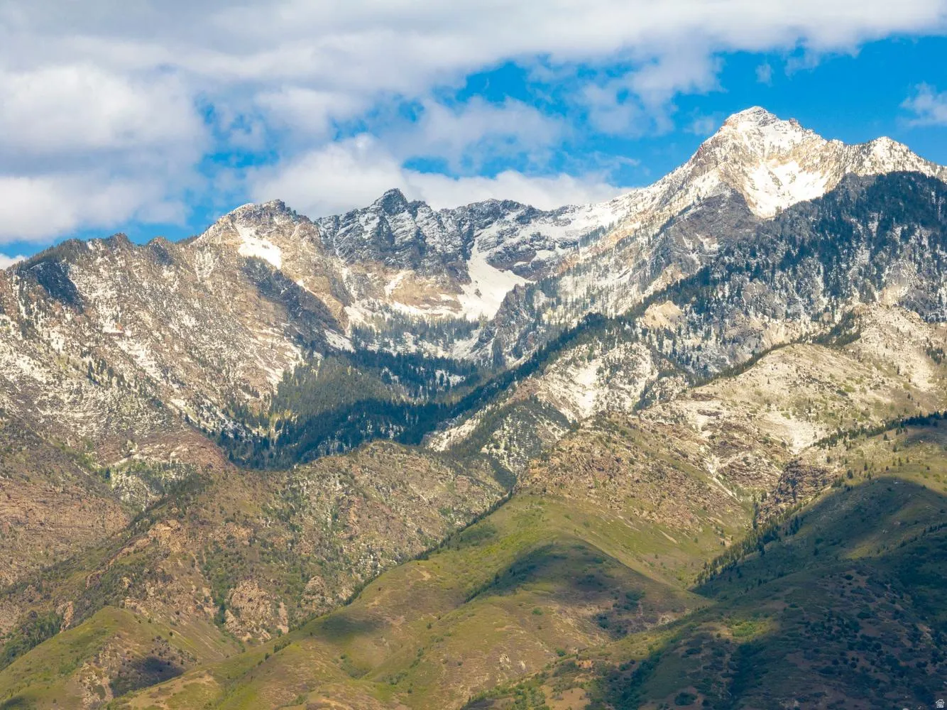 Mountain range featuring rugged peaks, snow-dusted slopes, varied green foliage, and a blue sky with scattered clouds