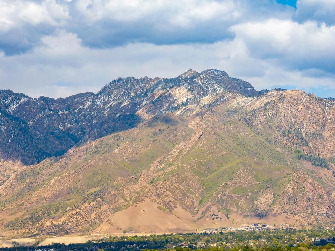 Mountain landscape featuring partially snow-capped peaks, varied green and brown foliage, and a residential area at the base