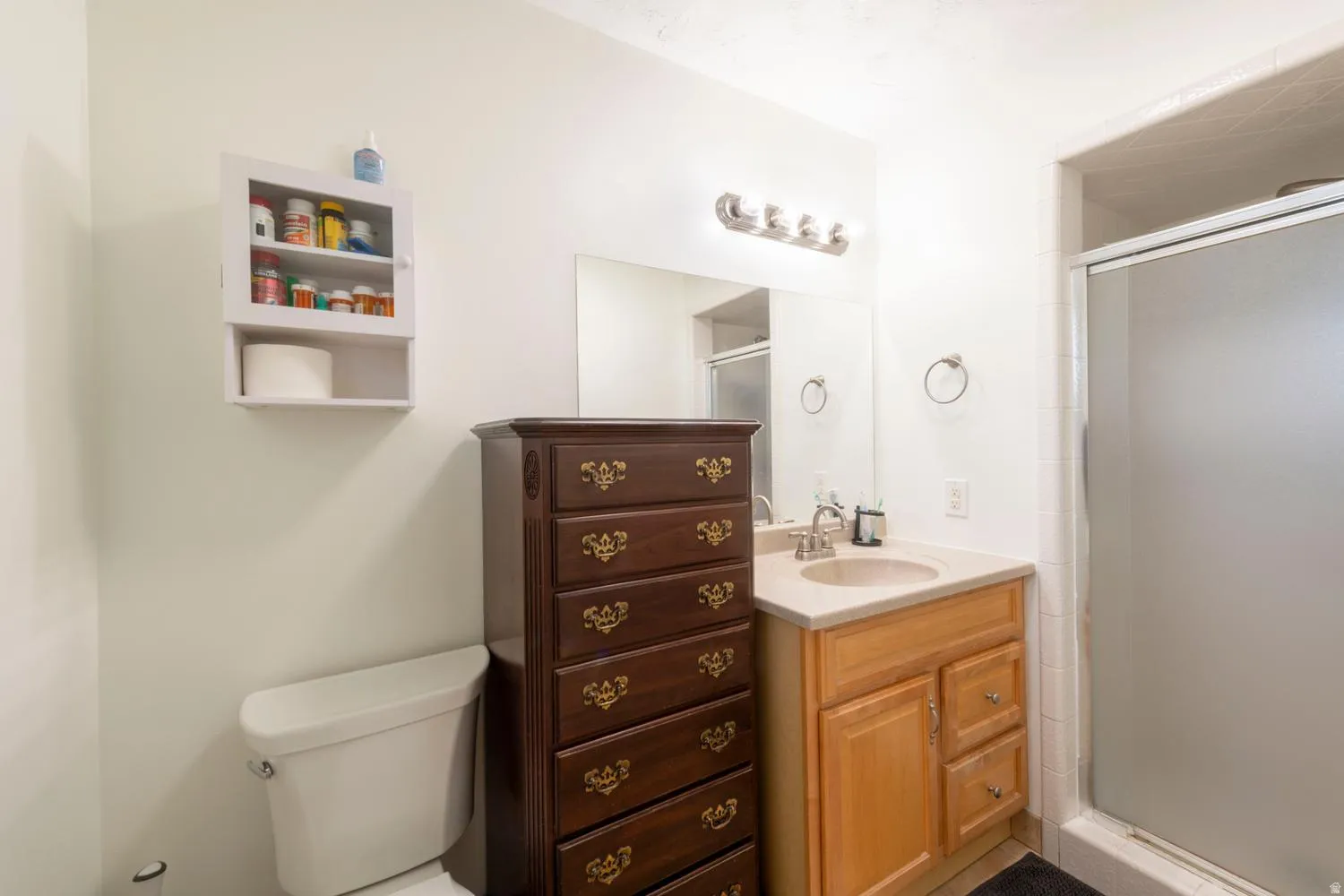 Bathroom featuring a single vanity with a light wood-finish cabinet and integrated sink, a large mirror, a four-bulb vanity light fixture, and a shower with a frosted sliding door
