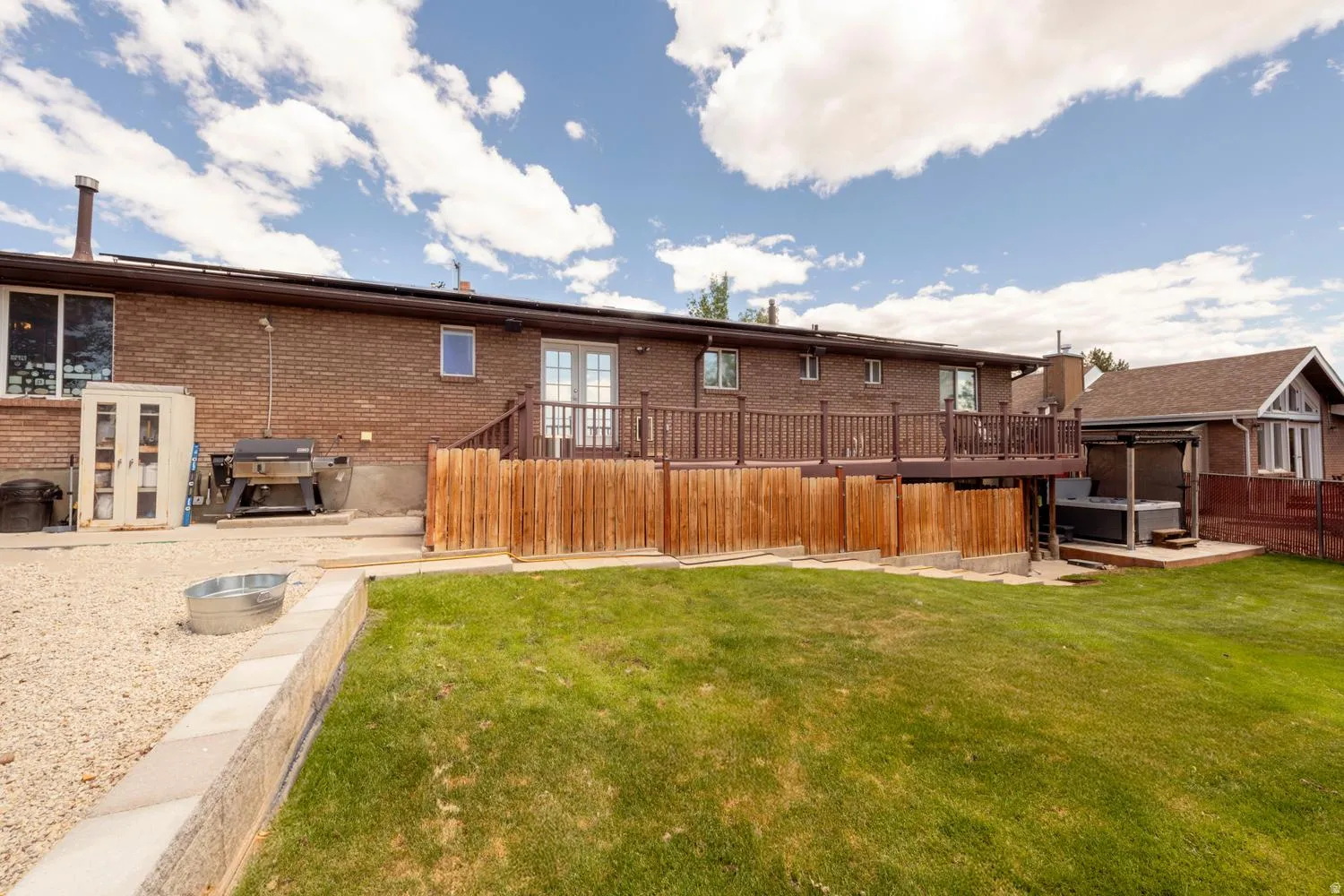 Spacious backyard featuring a manicured lawn, a raised wood deck with balusters, a stone retaining wall, and a hot tub with an overhead pergola