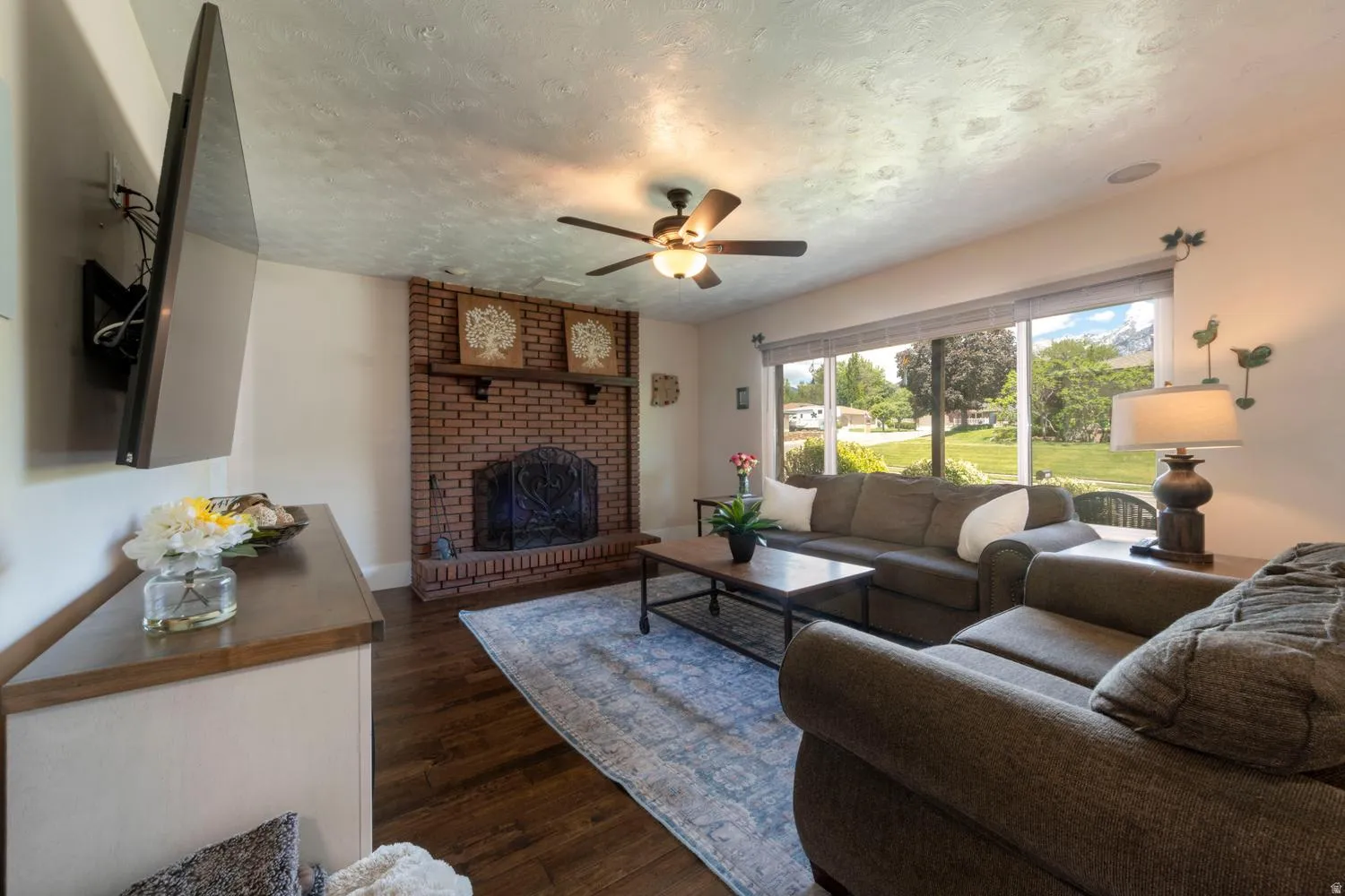 Spacious living area featuring expansive windows, wood-finish flooring, a brick fireplace with a mantel, and a ceiling fan