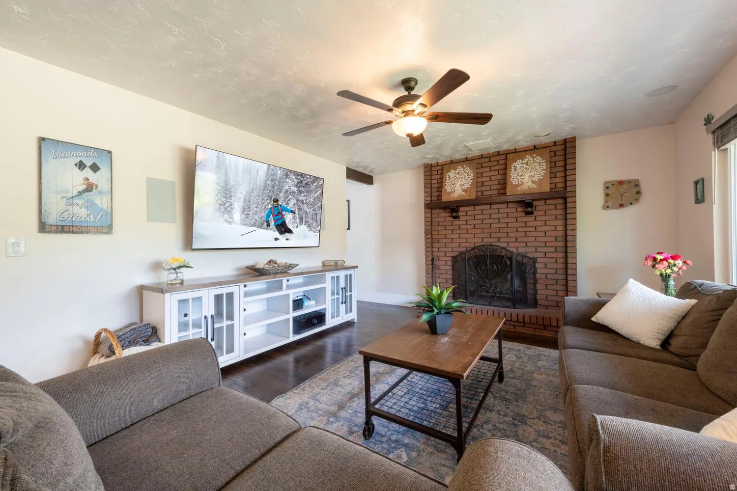Living area featuring a brick fireplace with a decorative mantel, dark wood-finish flooring, and a ceiling fan with integrated lighting