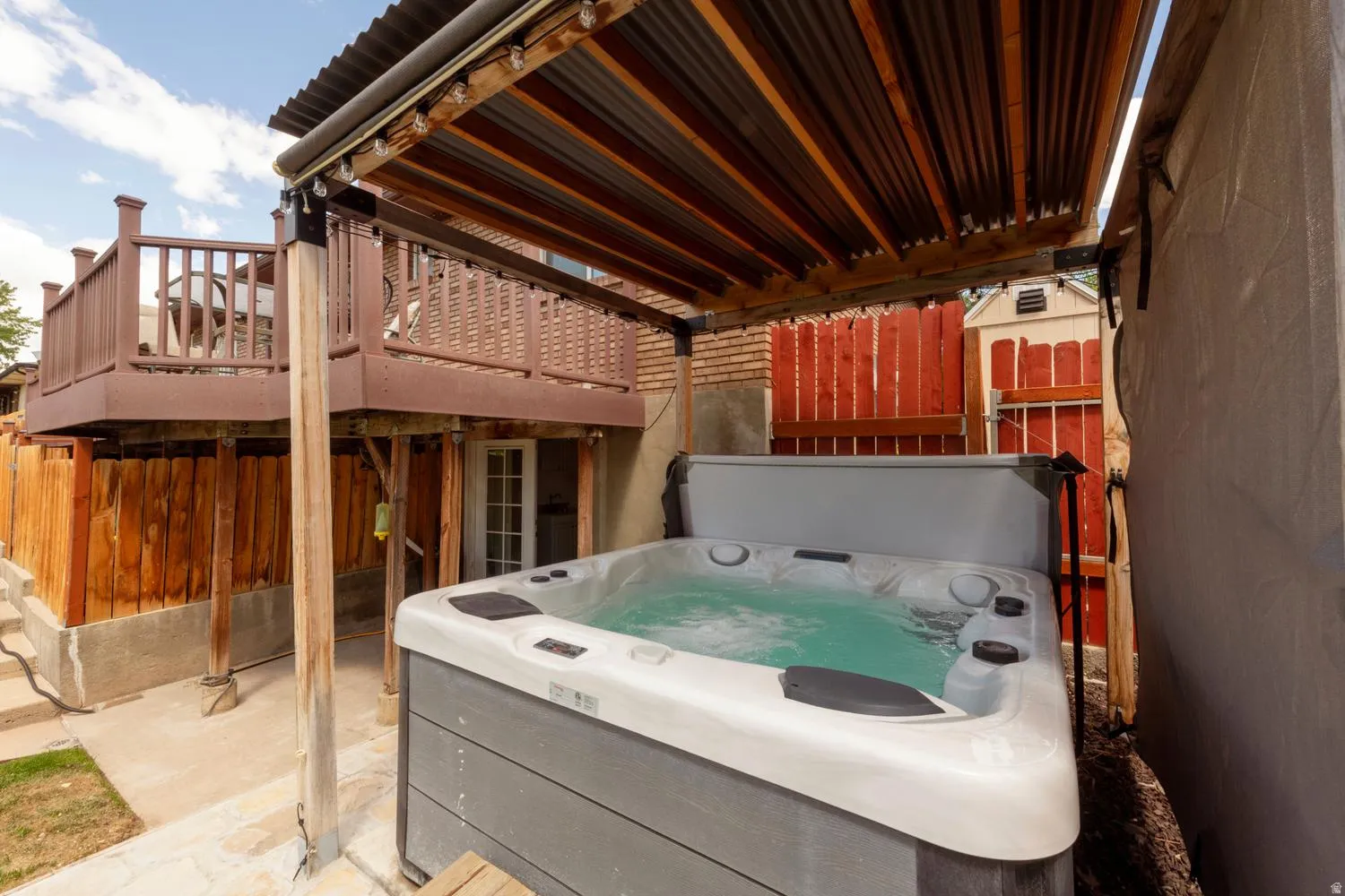 Outdoor spa with a corrugated metal and wood-slat overhead covering, surrounded by concrete paving