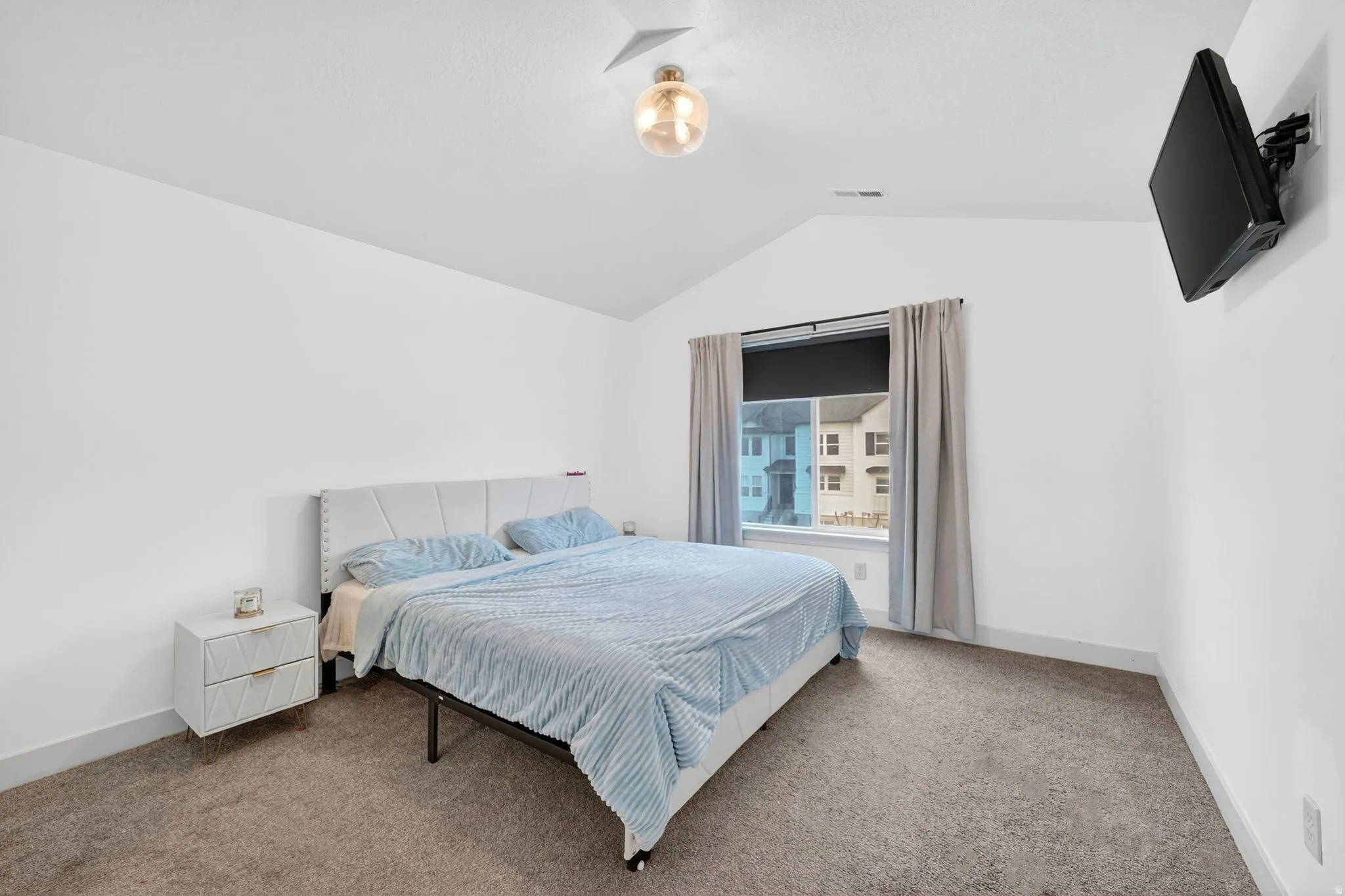 Bedroom featuring a vaulted ceiling with a contemporary light fixture, a large window with privacy blinds and drapes, and neutral-toned carpeting