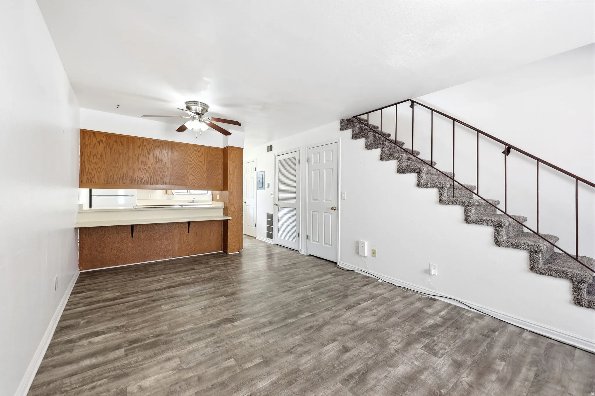 Spacious living area featuring wood-finish flooring, a carpeted staircase with a dark metal railing, and a kitchen pass-through with wood cabinetry